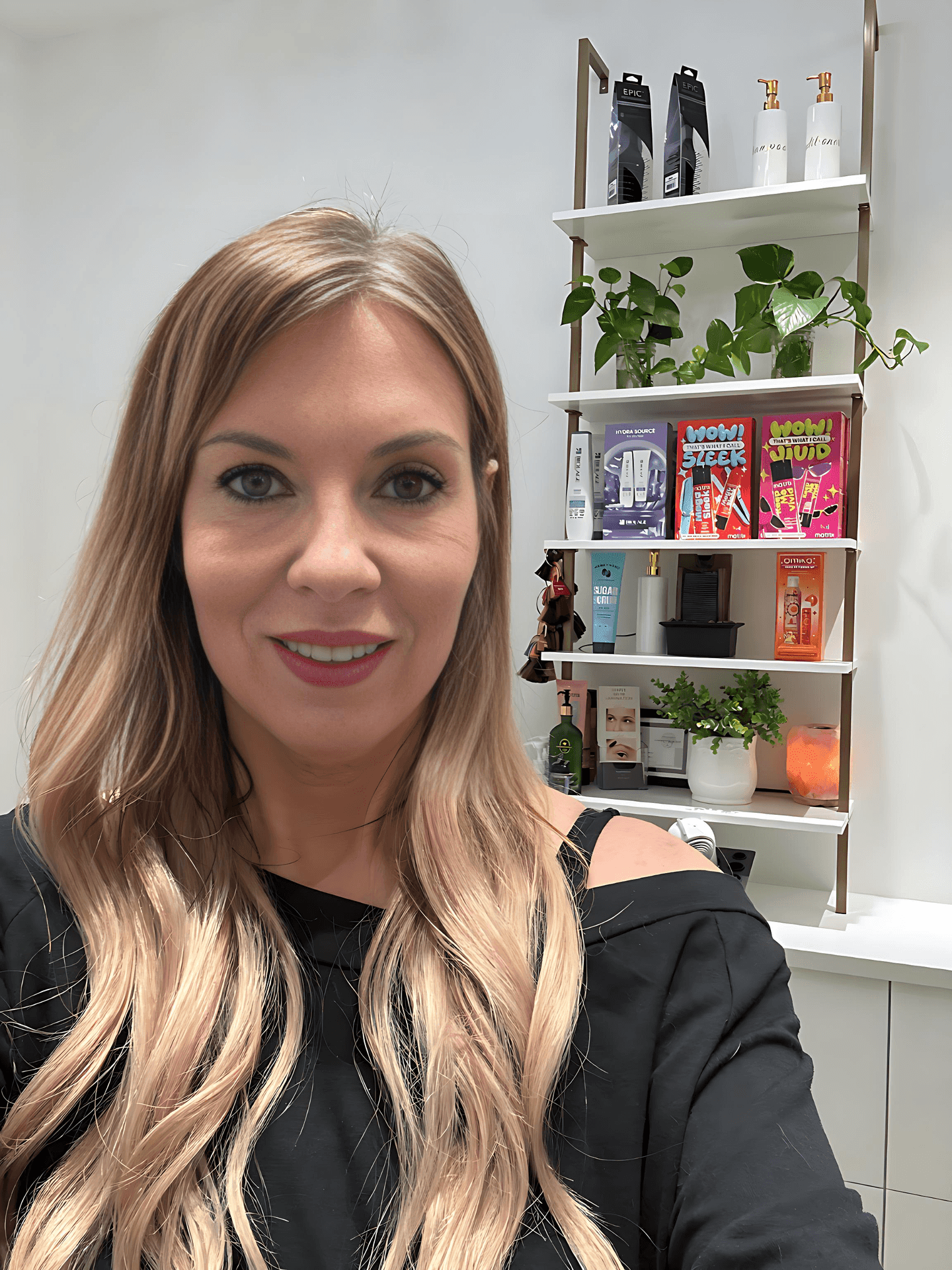 Woman with long hair posing in front of a shelf with beauty products and plants.